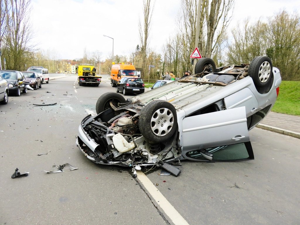 Overturned silver car on a road with debris, tow truck and responders in the background